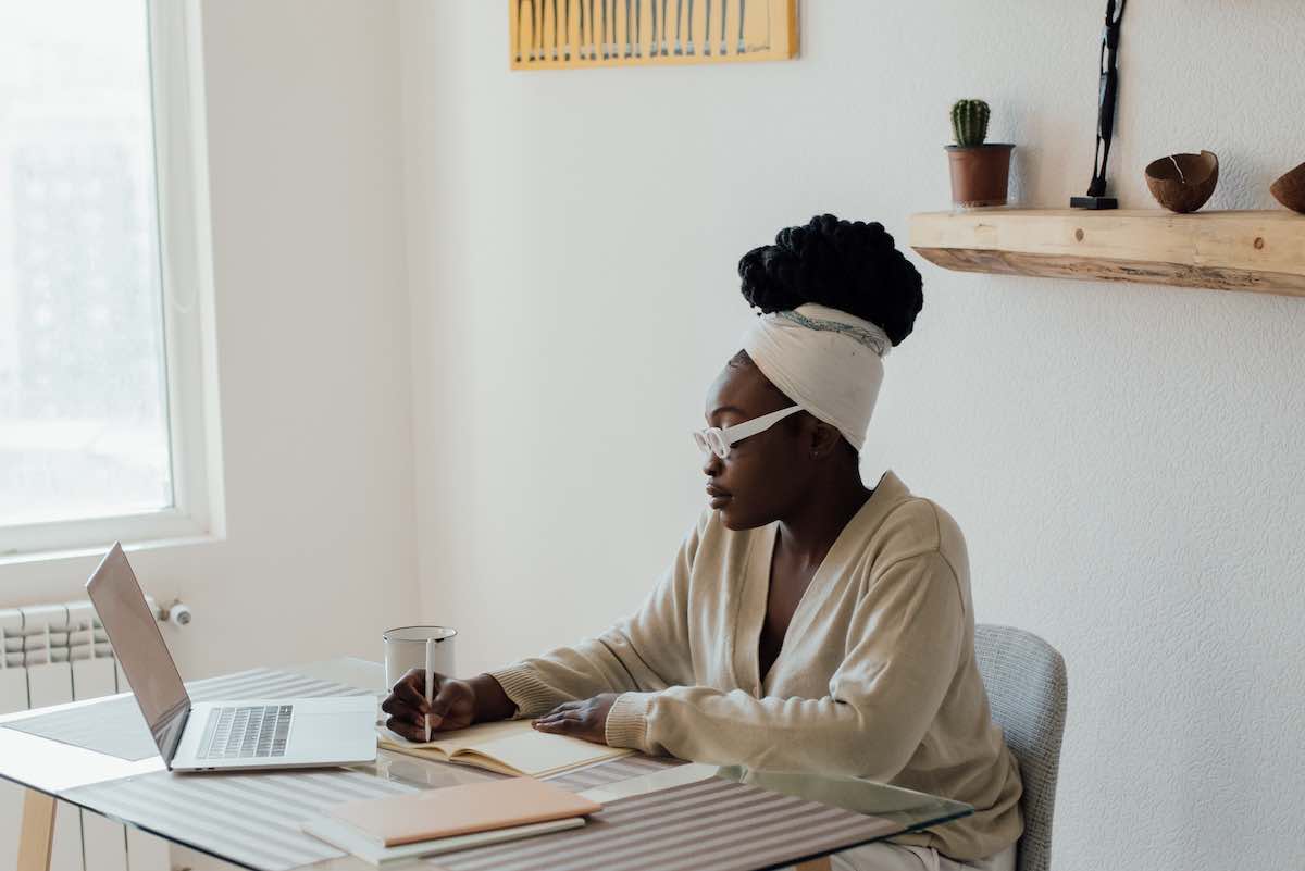 a woman writing on paper and sitting a desk with a laptop